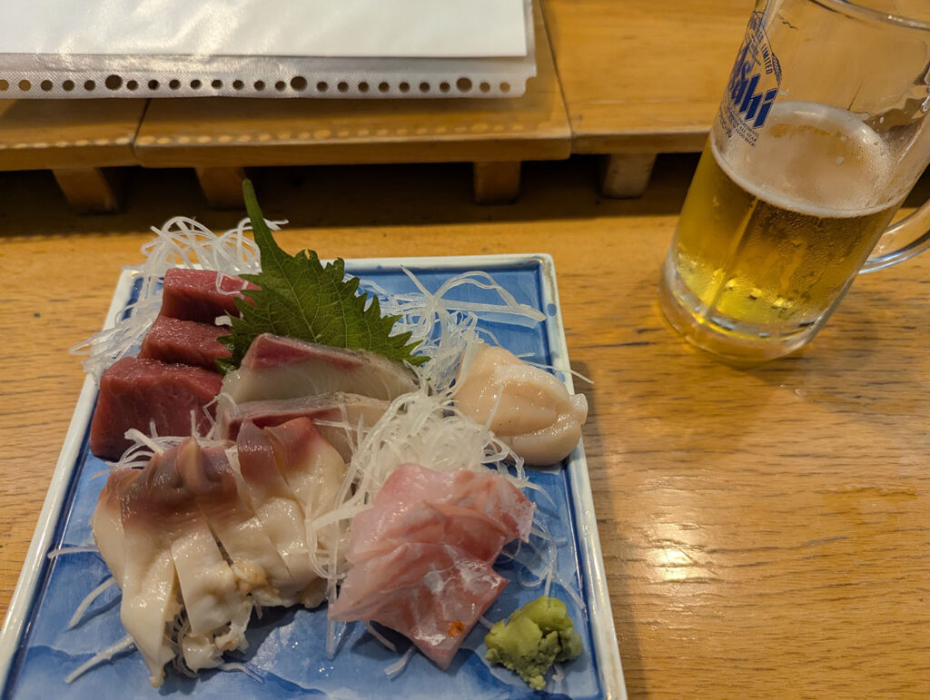木のカウンターに置かれた刺身盛り合わせとジョッキビール。新鮮な魚と冷えた一杯が並ぶ。