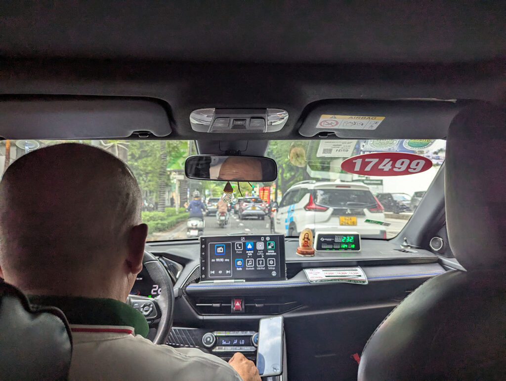 タクシーの後部座席から撮影した車内。運転手の後頭部、ダッシュボードのタブレット端末、フロントガラス越しに見えるバイクと車の流れ。メーターには「17499」の数字。