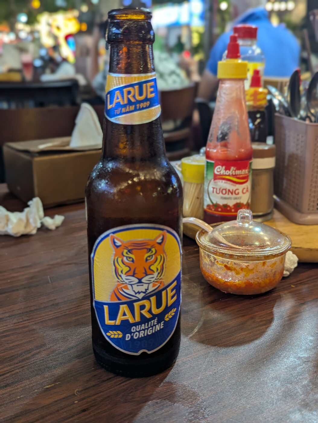 テーブルの上のLARUEビール1本と、ベトナムの調味料ボトル。奥にはティッシュの箱と夜の店内の灯り。