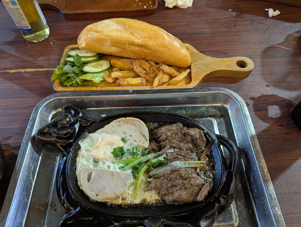 鉄板の上で焼かれたステーキ、目玉焼き、ベトナムソーセージ。横にはバインミーとフライドポテト。