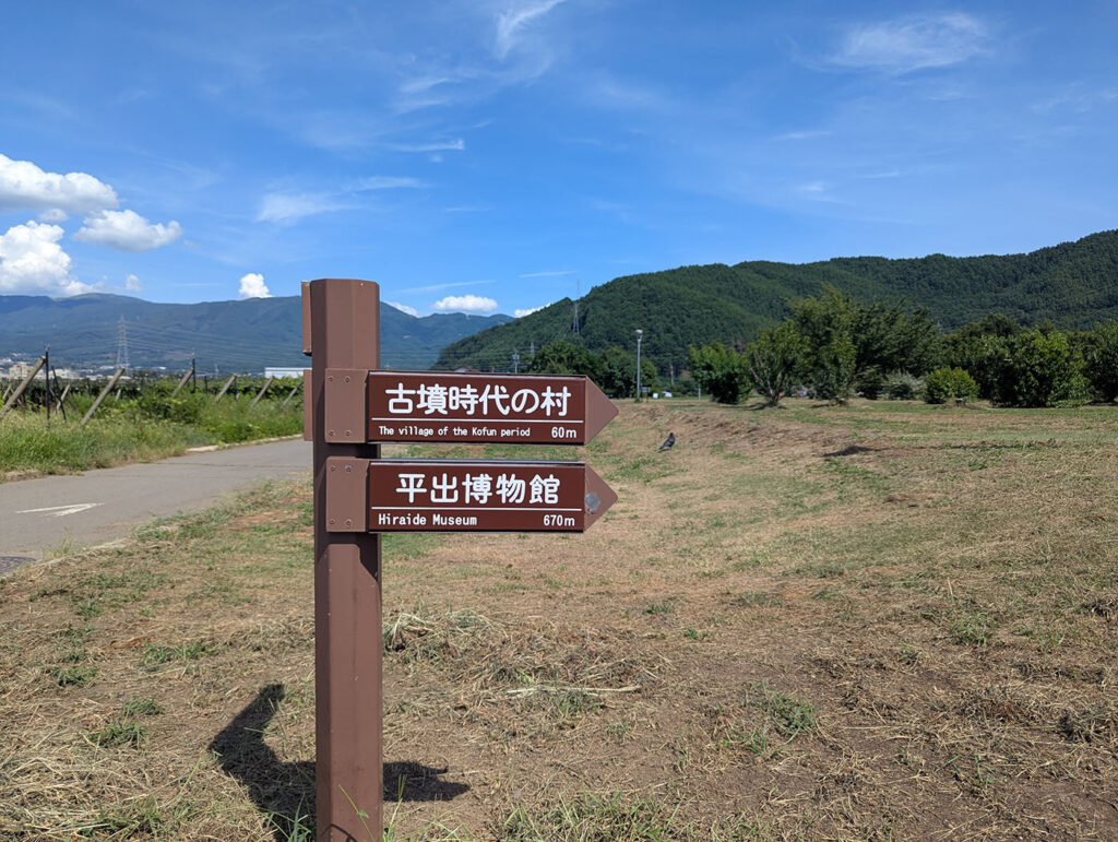 平出遺跡の案内標識（古墳時代の村・平出博物館への道）「古墳時代の村」「平出博物館」への距離を示す茶色の案内標識。背後には緑の山並みが広がり、のどかな風景が続く。