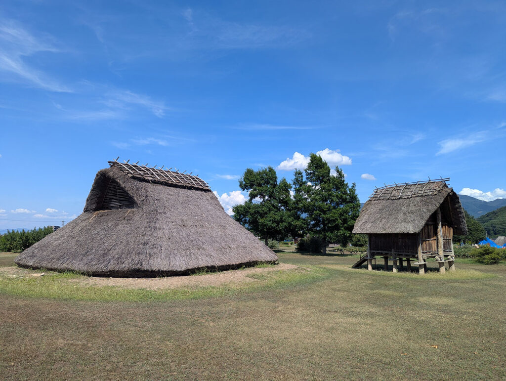 平出遺跡の復元竪穴住居と高床倉庫。広い草地に並ぶ竪穴住居と高床式倉庫。縄文から古墳時代の人々の暮らしを再現しており、青空の下に古代の風景がよみがえる。