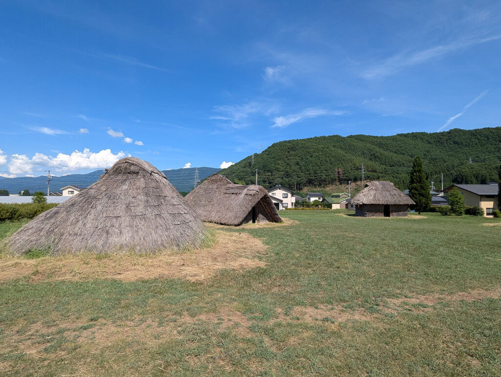 平出遺跡の竪穴住居群。竪穴住居が点在する平出遺跡の集落跡。背後には緑の山々が広がり、当時の自然と共に暮らした人々の営みを想像させる。